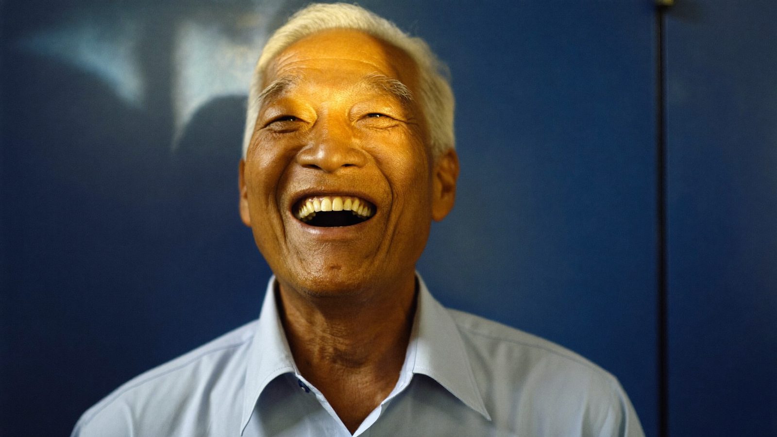 An older Singaporean man laughing with an open smile against a navy background