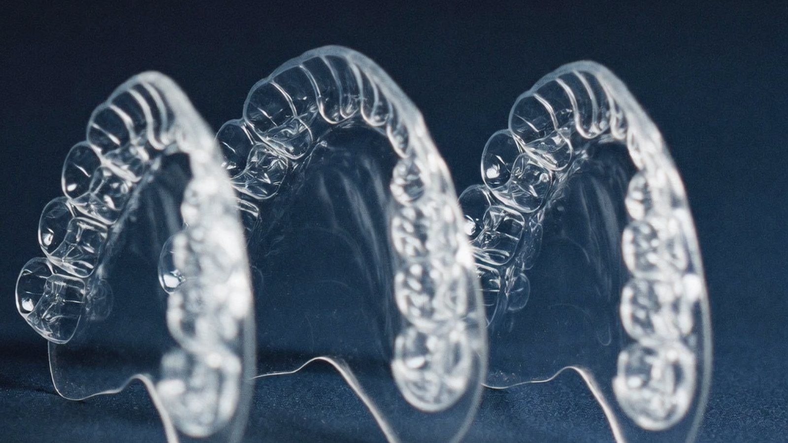 Row of clear orthodontic aligner trays receding into shallow focus on a deep navy surface